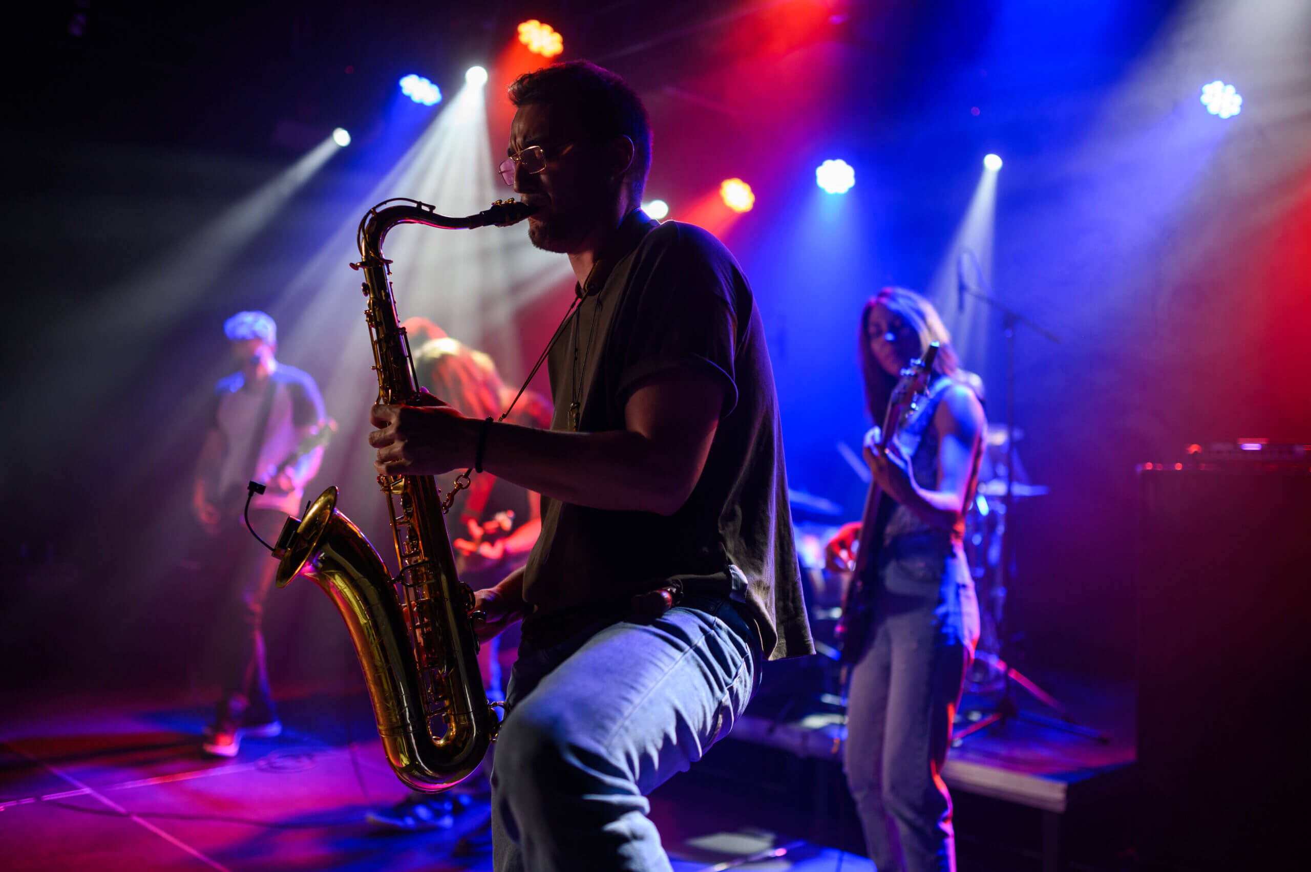 Saxophonist spielt auf einer Bühne mit bunten Lichtern und anderen Musikern im Hintergrund.